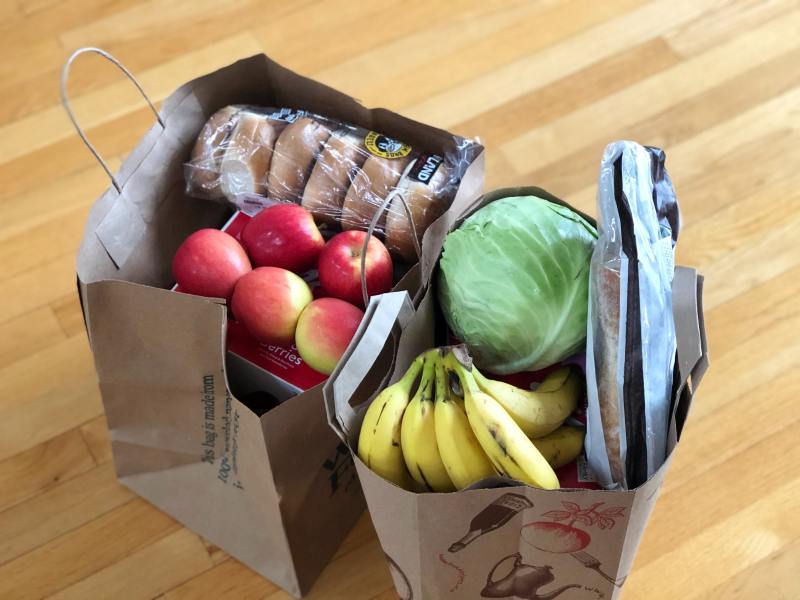 Two grocery bags sit on the floor of a hallway. They are stuffed full of apples, bananas, lettuce, and bread. 