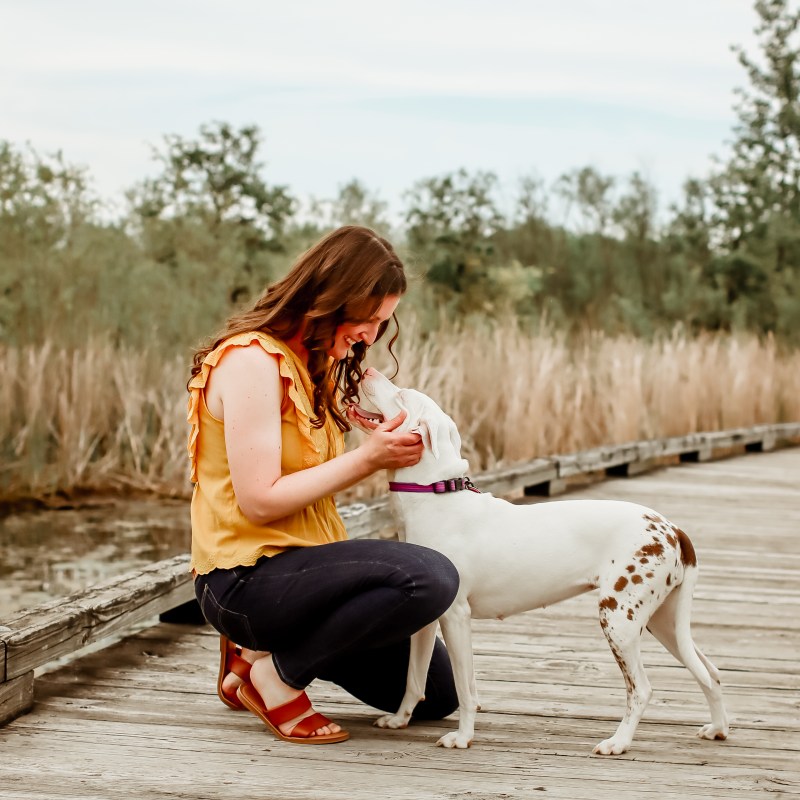 Me, a white woman with brown curled hair past my shoulders, wearing a yellow blouse and dark washed jeans, and my dog, Penny, a white dog with brown spots over her hips and tail. I'm crouched down giving her face some squishes. We're on a wood plank bridge with a marsh behind us. The day is overcast. 